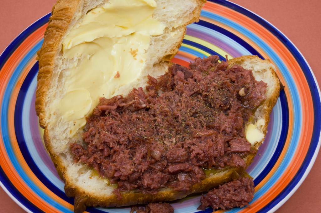 Open butter croissant sandwich on a colourful striped plate, topped with a generous layer of pan-fried hashed corned beef. Thick cultured butter is spread across the upper half of the flaky croissant and has begun to melt into the pastry. The corned beef filling is lightly browned and seasoned with black pepper, with a small amount spilling onto the plate. The image highlights the contrast between the buttery golden croissant, rich pink corned beef, and brightly coloured plate.