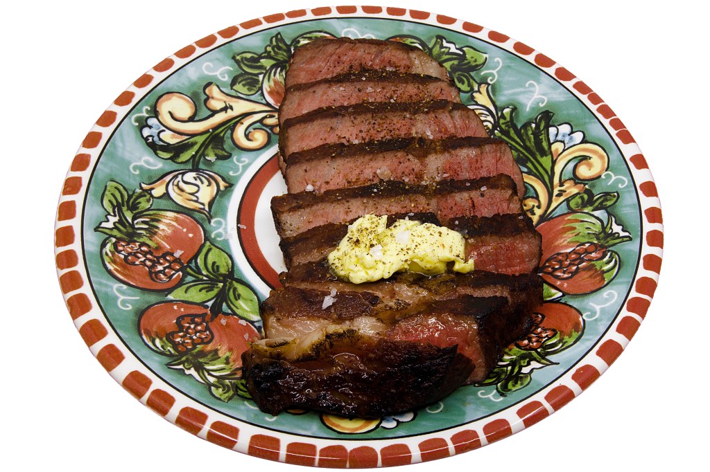 Sliced Scotch fillet steak arranged on a decorative ceramic plate, showing a deep brown seared crust and a pink medium-rare centre. The steak was cooked sous vide in a water bath and finished with a barbecue sear and gas torch, creating a charred exterior while retaining a juicy interior. A pat of butter rests on top of the sliced beef, beginning to melt over the warm meat, while flakes of coarse sea salt and cracked black pepper are scattered across the surface. The plate features colourful fruit and leaf motifs that contrast with the rich red and brown tones of the steak.