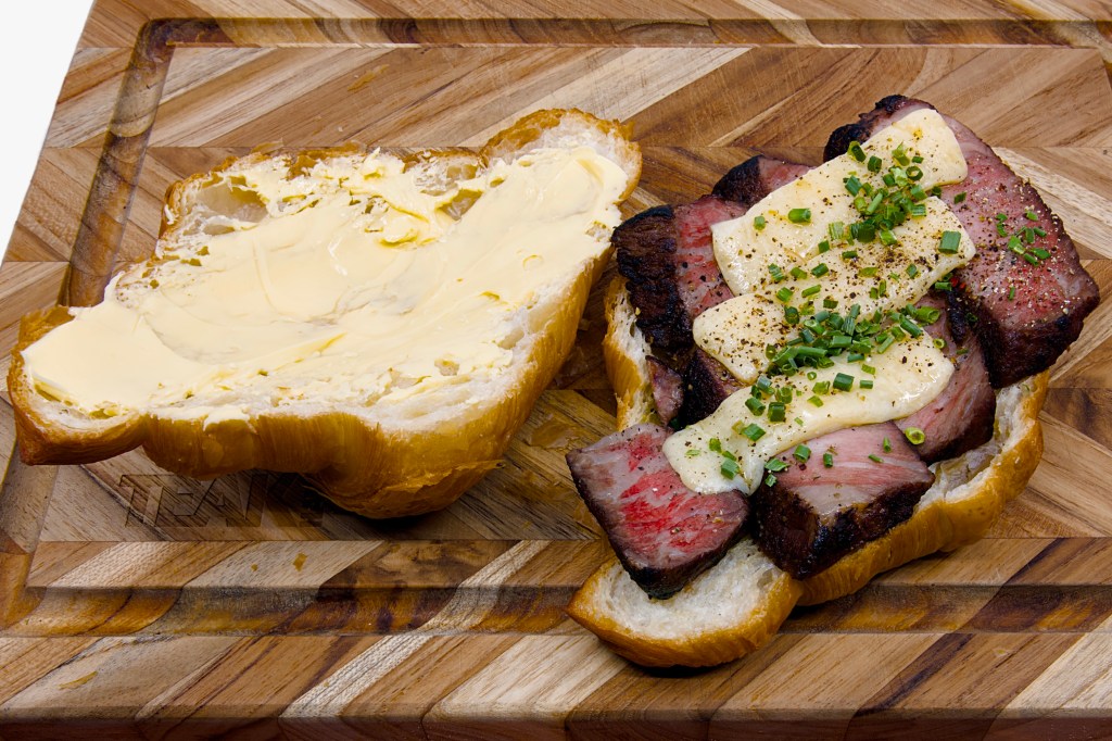Open toasted croissant sandwich on a wooden chopping board, filled with thick slices of leftover Scotch fillet steak and topped with melted vintage cheddar cheese. The croissant lid is generously spread with cultured butter, while the steak shows a dark, charred crust and a pink medium-rare centre. The melted cheese is sprinkled with cracked black pepper and finely chopped chives. The sandwich is presented partially assembled, highlighting the flaky croissant layers, buttery interior, and richly coloured steak and cheese filling.