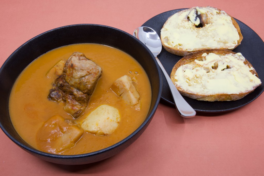 A hearty serving of beef short rib soup is presented in a matte black bowl. The smooth orange broth contains a large piece of braised beef short rib along with several chunks of tender potato. Beside the bowl, a black side plate holds two slices of (double-buttered) rye sourdough toast heavily spread with cultured butter, showing a thick layer of butter across the toasted crumb. A metal spoon rests between the soup and toast. The dark crockery contrasts with the creamy butter and vibrant soup, while the pink background gives the image a warm and inviting appearance.