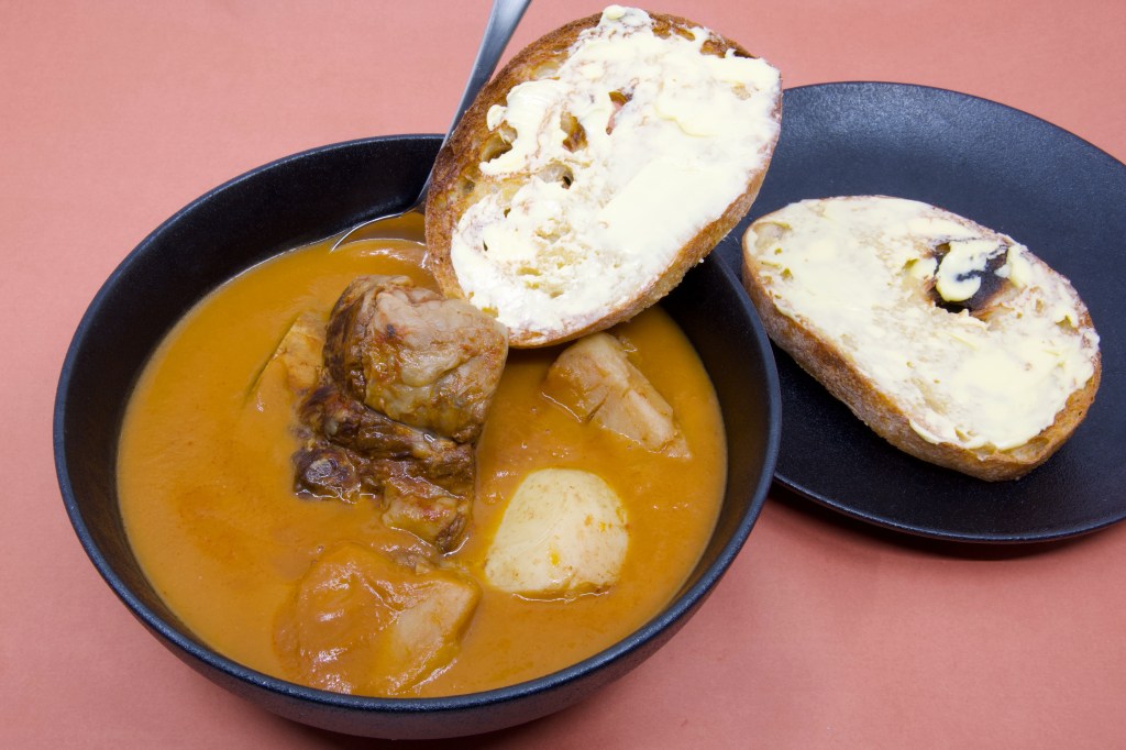 A dark bowl filled with beef short rib soup sits on a pink background. The smooth orange broth contains one piece of slow-cooked beef short rib and pieces of soft potato. A metal spoon is tucked behind a slice of double-buttered rye sourdough toast resting across the rim of the bowl. The sourdough is heavily spread with cultured butter, and a second buttered slice sits on a matching black plate beside the bowl. The contrast between the dark crockery, creamy butter, and warm-coloured soup creates a hearty and inviting presentation.