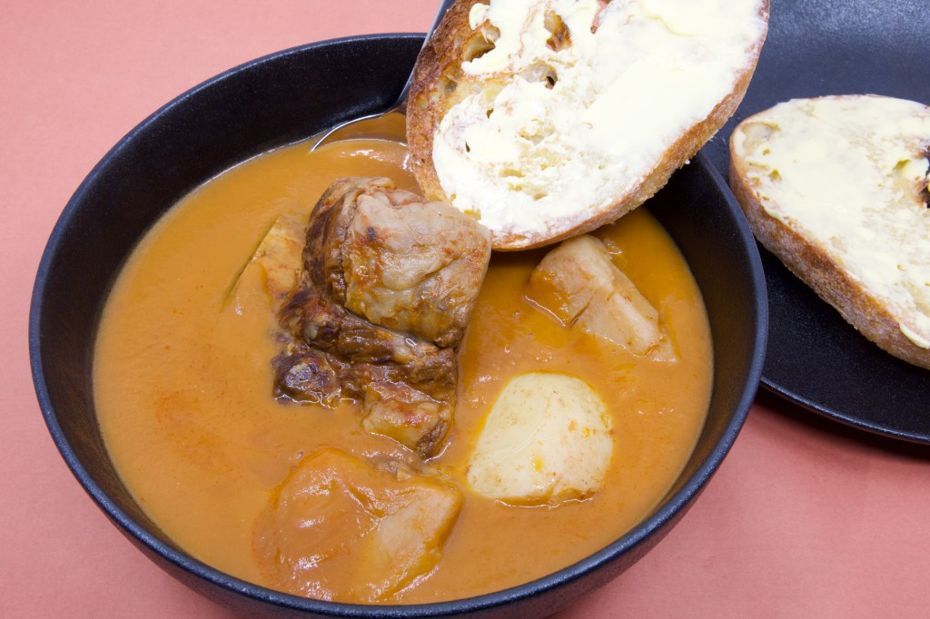 A dark bowl filled with beef short rib soup sits on a pink background. The smooth orange broth contains one piece of slow-cooked beef short rib and pieces of soft potato. A metal spoon is tucked behind a slice of double-buttered rye sourdough toast resting across the rim of the bowl. The sourdough is heavily spread with cultured butter, and a second buttered slice sits on a matching black plate beside the bowl. The contrast between the dark crockery, creamy butter, and warm-coloured soup creates a hearty and inviting presentation.