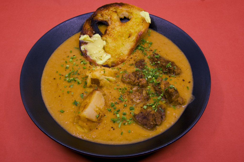 A bowl of pressure-cooked beef short rib sits in a rich, velvety broth puréed from the cooking liquor, fennel, brown onion, celery, and potato. Several chunks of beef and potato are visible above the surface, scattered with freshly chopped chives and cracked black pepper. Resting against the rim of the bowl are two thick slices of double-butted sourdough toast, generously spread with melting butter. The black bowl is photographed against a red background, highlighting the warm golden tones of the broth.