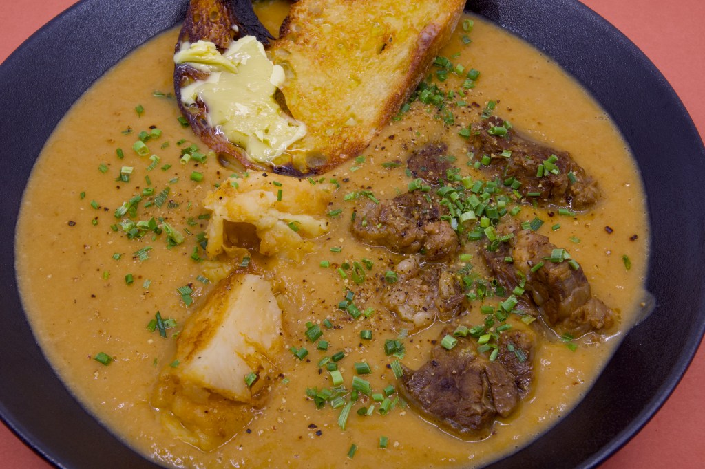 A bowl of pressure-cooked beef short rib sits in a rich, velvety broth puréed from the cooking liquor, fennel, brown onion, celery, and potato. Several chunks of beef and potato are visible above the surface, scattered with freshly chopped chives and cracked black pepper. Resting against the rim of the bowl are two thick slices of double-butted sourdough toast, generously spread with melting butter. The black bowl is photographed against a red background, highlighting the warm golden tones of the broth.