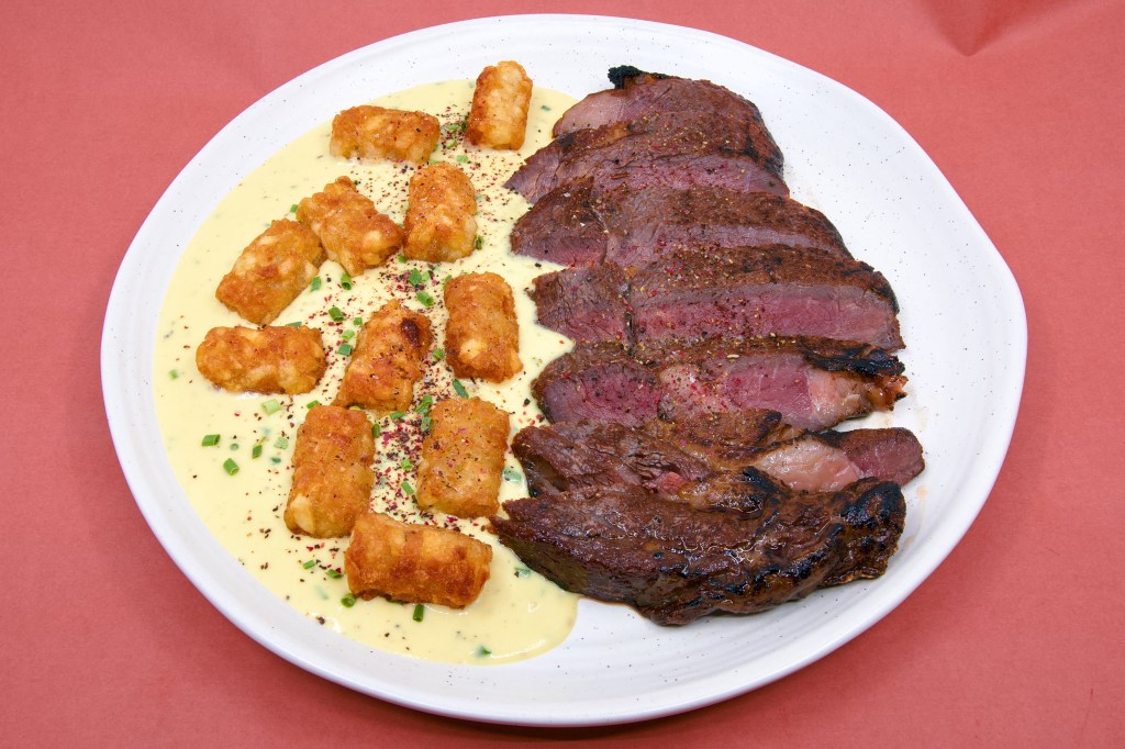 Close overhead photograph of dry-brined scotch fillet steak cooked sous vide at 57 °C and seared with a gas blow torch. Golden potato gems sit on a velvety blue cheese sauce made with cultured butter, cream, cream cheese, blue brie cheese, and Dijon mustard, topped with peppercorns and chopped chives.