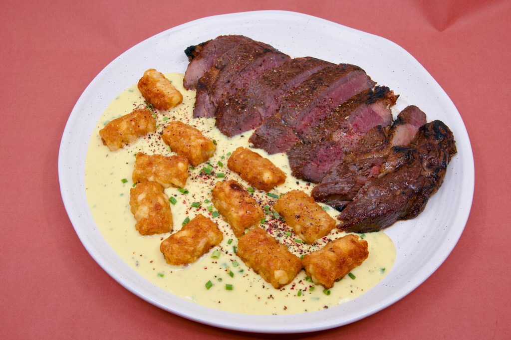 Overhead view of medium-rare scotch fillet steak slices with a charred crust from gas blow-torch searing. Beef-fat potato gems rest on a smooth blue cheese sauce, with freshly cracked peppercorns and chopped chives scattered across the plate.