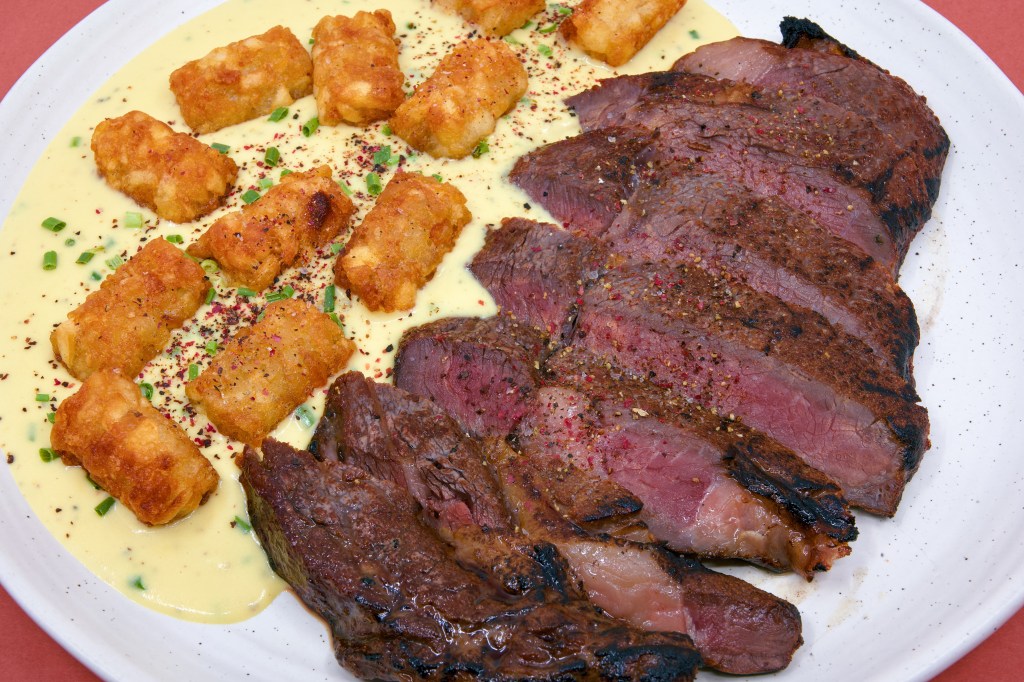 Plate of sliced scotch fillet steak cooked sous vide and torch-seared, accompanied by golden potato gems cooked in beef fat. The dish sits on a rich blue cheese sauce made with cultured butter, cream, cream cheese, blue brie, and Dijon mustard, garnished with cracked pepper and fresh chives.