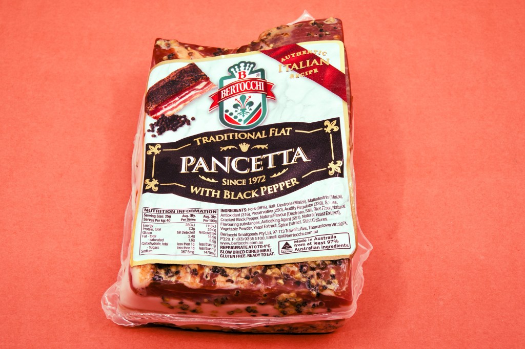 A vacuum-packed slab of Bertocchi Traditional Flat Pancetta with Black Pepper sits on a coral pink background. The cured pork belly is coated with cracked black pepper, and the front label highlights the Italian-style pancetta ready for cooking or serving.