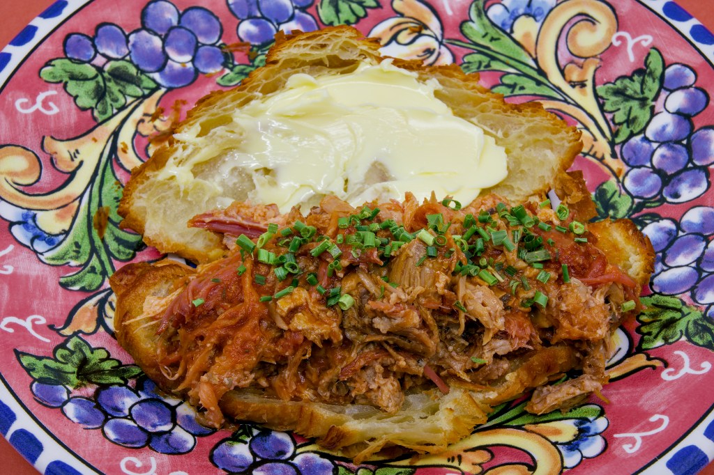 Open croissant sandwich filled with juicy pulled pork made from pork ribs and pancetta. The flaky croissant is generously spread with cultured butter and topped with chopped chives, all presented on a brightly patterned ceramic plate.