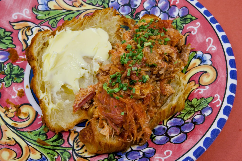Open croissant sandwich filled with juicy pulled pork made from pork ribs and pancetta. The flaky croissant is generously spread with cultured butter and topped with chopped chives, all presented on a brightly patterned ceramic plate.