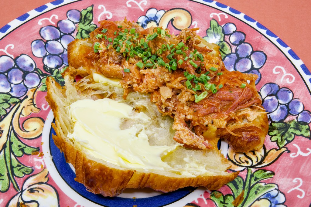 Open croissant sandwich filled with juicy pulled pork made from pork ribs and pancetta. The flaky croissant is generously spread with cultured butter and topped with chopped chives, all presented on a brightly patterned ceramic plate.