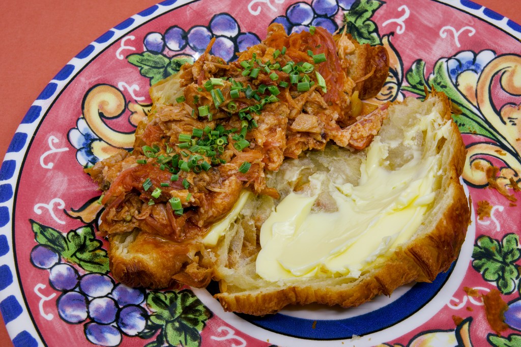 Open croissant sandwich filled with juicy pulled pork made from pork ribs and pancetta. The flaky croissant is generously spread with cultured butter and topped with chopped chives, all presented on a brightly patterned ceramic plate.