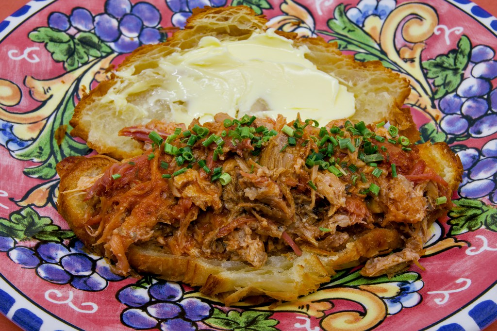 Open croissant sandwich filled with juicy pulled pork made from pork ribs and pancetta. The flaky croissant is generously spread with cultured butter and topped with chopped chives, all presented on a brightly patterned ceramic plate.
