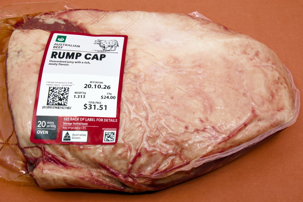 A whole beef rump cap in vacuum-sealed plastic packaging sits on a brown bench or tabletop. The large triangular cut is covered by a thick layer of creamy-white fat, with dark red beef visible around the edges. A supermarket label on the front identifies the cut as Australian beef rump cap, weighing 1.313 kilograms. The image shows the roast in its original retail packaging before preparation and cooking.