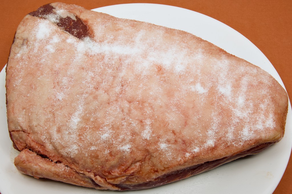 A whole beef rump cap sits on a large white plate atop a brown benchtop. The creamy-white fat cap has been heavily sprinkled with coarse salt, creating a visible crystalline coating across the surface. The triangular cut is shown fat-side up and is ready for an overnight dry brine, a process that helps season the meat and improve browning and flavour before cooking.