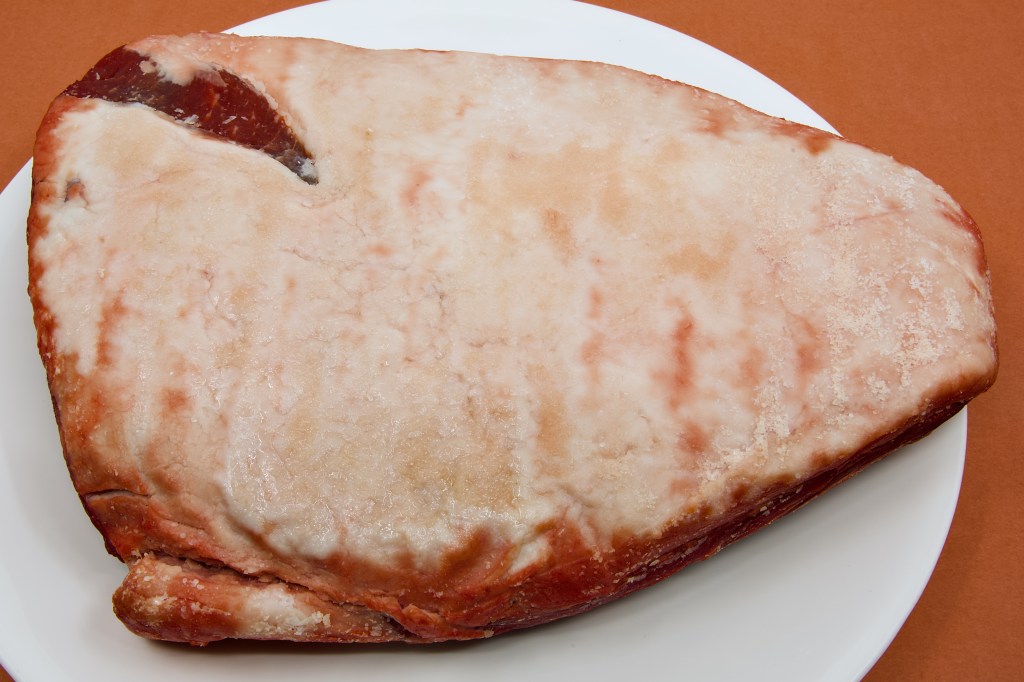 A whole beef rump cap sits fat-side up on a large white plate after being dry brined overnight. The coarse salt applied the previous day has largely dissolved, drawing moisture to the surface and creating a slightly glossy sheen across the creamy-white fat cap. Only small patches of salt crystals remain visible. The roast is shown prepared and ready for cooking, with the deep red beef visible around the edges beneath the fat layer.