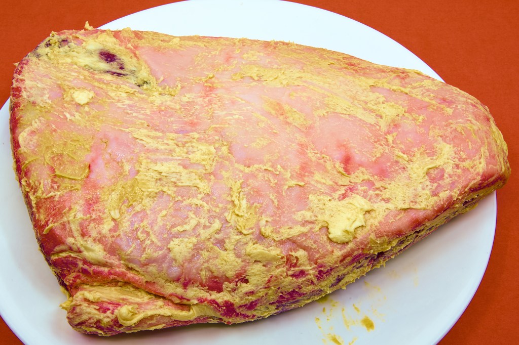 A whole beef rump cap sits on a large white plate on a brown benchtop. Following an overnight dry brine, the thick fat cap has been rubbed with a mixture of yellow mustard and butter, leaving a pale golden coating across the surface. The triangular roast remains fat-side up, with some dark red beef visible around the edges. The mustard and butter are spread in a rustic layer, preparing the roast for oven cooking and the development of a richly browned crust.