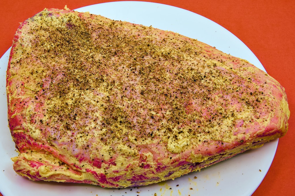 A whole beef rump cap sits fat-side up on a large white plate on a brown benchtop. After an overnight dry brine, the fat cap has been rubbed with mustard and butter, creating a pale golden coating over the surface. Freshly ground black pepper and sea salt have been sprinkled generously across the roast, forming a speckled seasoning layer. The triangular cut is ready for roasting, with the rich red beef visible around the edges beneath the thick fat cap.