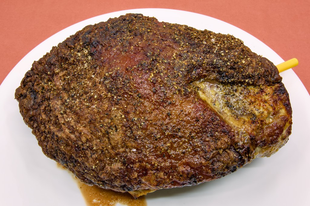 A roasted beef rump cap sits on a large white plate against a light brown background. The surface of the roast is covered with a richly browned crust formed from mustard, butter, salt, and black pepper. Rendered fat has caramelised across the fat cap, creating a glossy finish with dark golden-brown and mahogany tones. A yellow Combustion Inc wireless thermometer remains inserted into the roast to monitor the internal temperature during the resting period. Small amounts of meat juice and rendered fat have collected on the plate beneath the roast, highlighting the moist, freshly cooked appearance of the beef.