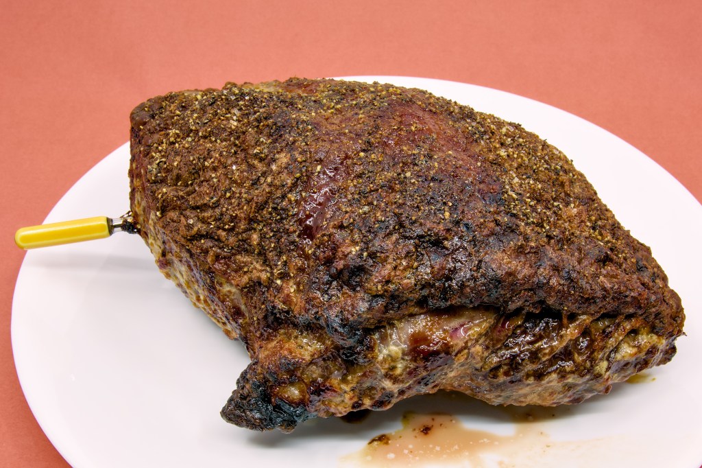 A roasted beef rump cap sits on a large white plate against a light brown background. The surface of the roast is covered with a richly browned crust formed from mustard, butter, salt, and black pepper. Rendered fat has caramelised across the fat cap, creating a glossy finish with dark golden-brown and mahogany tones. A yellow Combustion Inc wireless thermometer remains inserted into the roast to monitor the internal temperature during the resting period. Small amounts of meat juice and rendered fat have collected on the plate beneath the roast, highlighting the moist, freshly cooked appearance of the beef.