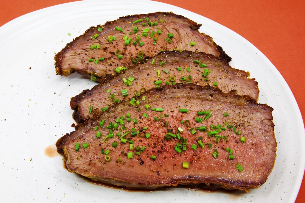 Three generous slices of roast beef rump cap are served on a large white plate against a pale brown background. Each slice reveals a moist, blush-pink interior framed by a thin layer of rendered fat and a well-browned crust seasoned with black pepper. The beef is garnished with freshly cracked peppercorns and finely chopped chives, which add black and bright green flecks across the surface. A small pool of meat juices has collected beneath the slices, highlighting the tenderness and moisture of the roast. No vegetables, sauce, or other accompaniments are served with the beef.