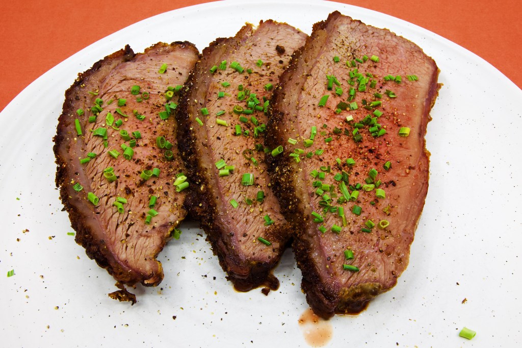 Three generous slices of roast beef rump cap are served on a large white plate against a pale brown background. Each slice reveals a moist, blush-pink interior framed by a thin layer of rendered fat and a well-browned crust seasoned with black pepper. The beef is garnished with freshly cracked peppercorns and finely chopped chives, which add black and bright green flecks across the surface. A small pool of meat juices has collected beneath the slices, highlighting the tenderness and moisture of the roast. No vegetables, sauce, or other accompaniments are served with the beef.
