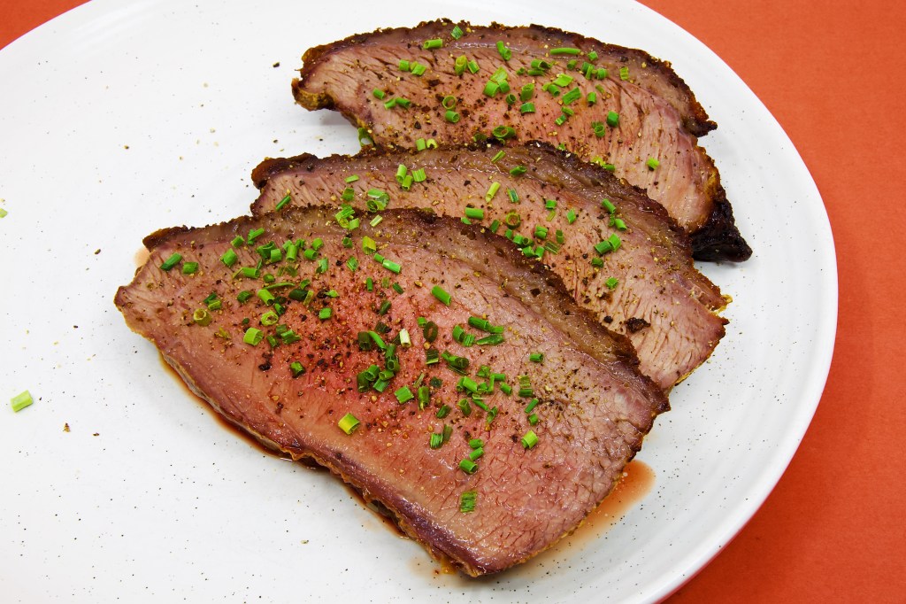 Three generous slices of roast beef rump cap are served on a large white plate against a pale brown background. Each slice reveals a moist, blush-pink interior framed by a thin layer of rendered fat and a well-browned crust seasoned with black pepper. The beef is garnished with freshly cracked peppercorns and finely chopped chives, which add black and bright green flecks across the surface. A small pool of meat juices has collected beneath the slices, highlighting the tenderness and moisture of the roast. No vegetables, sauce, or other accompaniments are served with the beef.