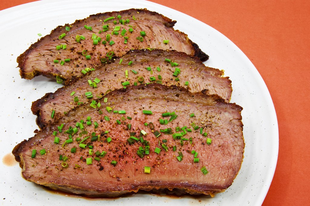 Three generous slices of roast beef rump cap are served on a large white plate against a pale brown background. Each slice reveals a moist, blush-pink interior framed by a thin layer of rendered fat and a well-browned crust seasoned with black pepper. The beef is garnished with freshly cracked peppercorns and finely chopped chives, which add black and bright green flecks across the surface. A small pool of meat juices has collected beneath the slices, highlighting the tenderness and moisture of the roast. No vegetables, sauce, or other accompaniments are served with the beef.