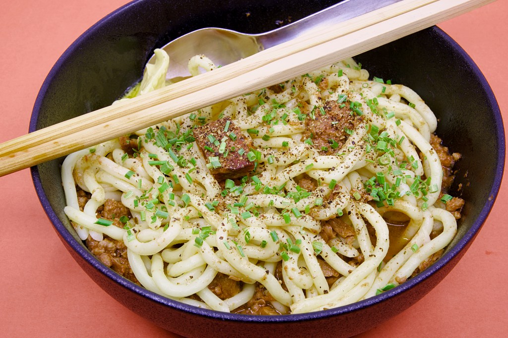 Bowl of Hokkien noodles topped with mixed mammal stew made from beef, lamb, and pork, finished with chopped chives, cracked black pepper, butter, chopsticks, and a spoon.