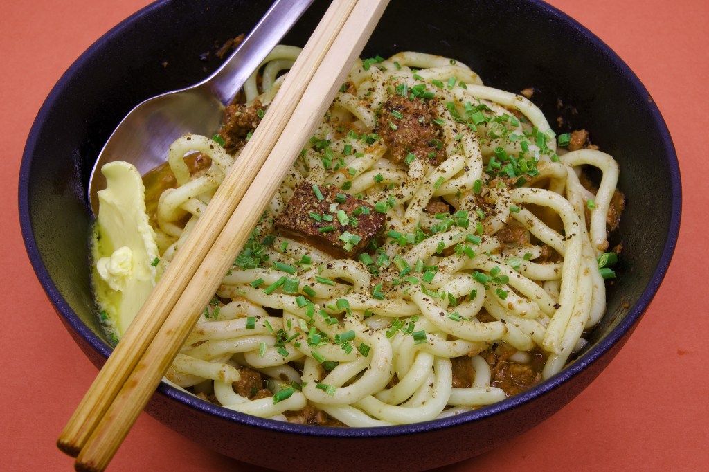 Bowl of Hokkien noodles topped with mixed mammal stew made from beef, lamb, and pork, finished with chopped chives, cracked black pepper, butter, chopsticks, and a spoon.