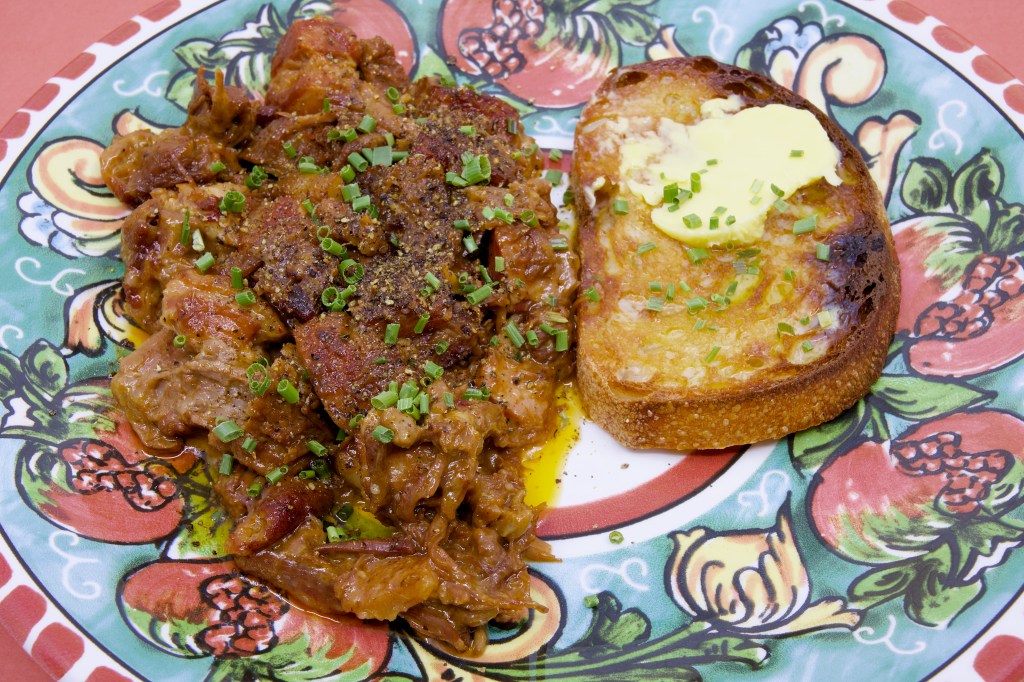 Mixed mammal stew made with beef chuck, lamb leg, and pork belly served on a decorative plate beside a slice of double-buttered rye sourdough toast topped with melted butter and chives.