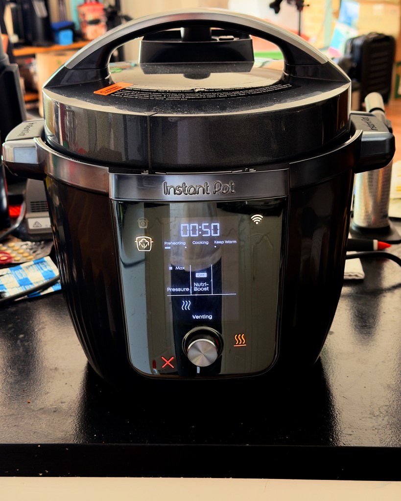 A black Instant Pot pressure cooker sits on a kitchen benchtop with the digital display lit. The screen shows a 50-minute cooking time, pressure-cooking settings, and venting mode, highlighting the appliance used to prepare a slow-cooked pork and pancetta dish.