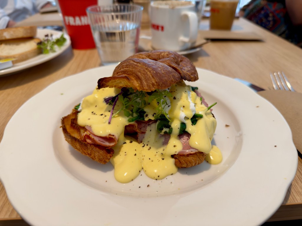 Eggs Benedict served on a large buttery croissant, filled with folded ham, poached eggs, baby greens, and generously covered in creamy hollandaise sauce. The dish sits on a white plate at a café table at Frankie's on Franklin, with coffee cups and water glasses softly blurred in the background.