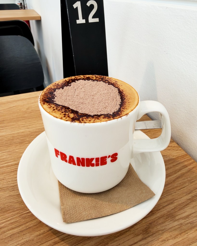 A large mug of full-cream milk cappuccino at Frankie’s on Franklin, topped with chocolate powder and served on a white saucer beside table 12.