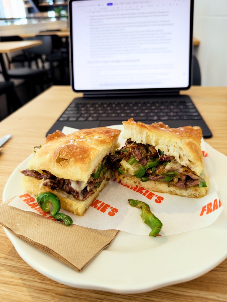Cheesesteak focaccia sandwich cut in half on a white plate at Frankie’s on Franklin, showing sliced beef, melted cheese, green capsicum, and onion, with a laptop on the table behind