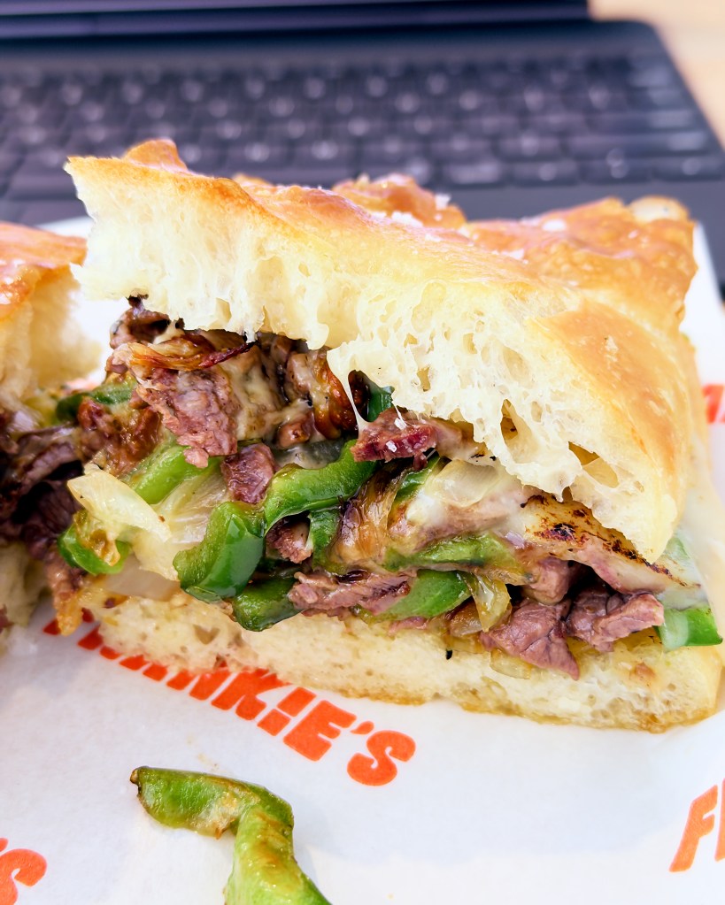 Detailed close-up of a cheesesteak focaccia sandwich showing the airy focaccia crumb, sliced beef, melted cheese, sautéed onion, and green capsicum filling.