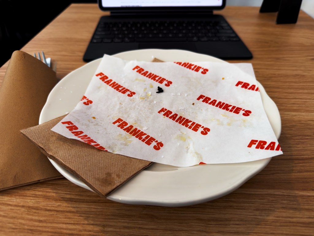 Empty white plate lined with Frankie’s branded paper and napkins after finishing a cheesesteak focaccia sandwich, with a laptop visible in the background.